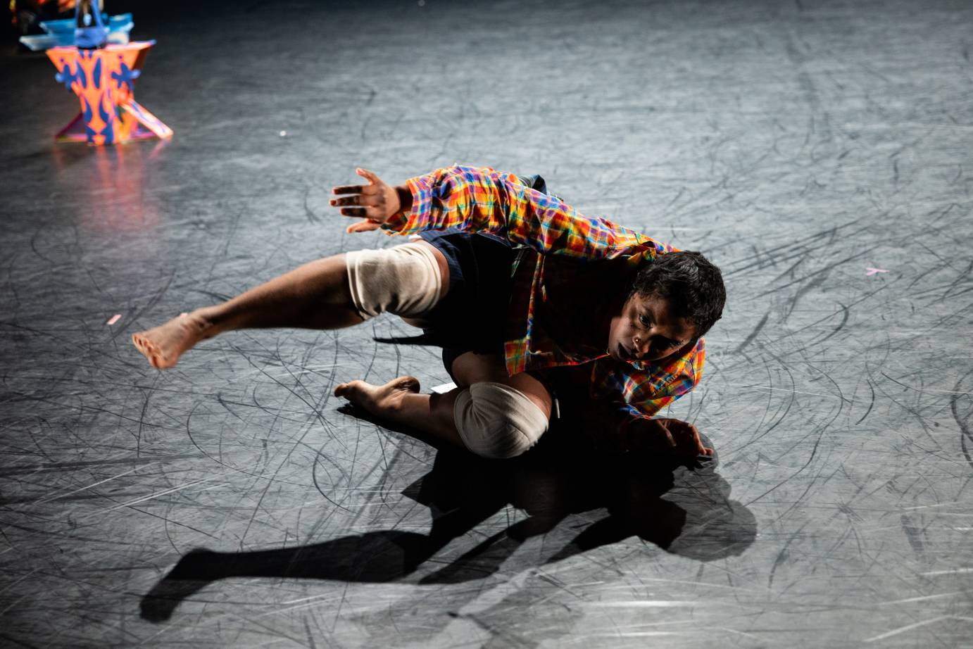 a woman in a colorful flannel shirt, denim shorts and knee pads appears to skid on the stage along the side of her body