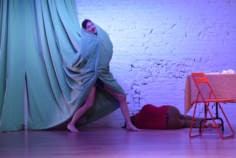 Gabrielle wraps herself in a sage-colored curtain. Aleksandr lays on the ground. His back faces away from the audience.