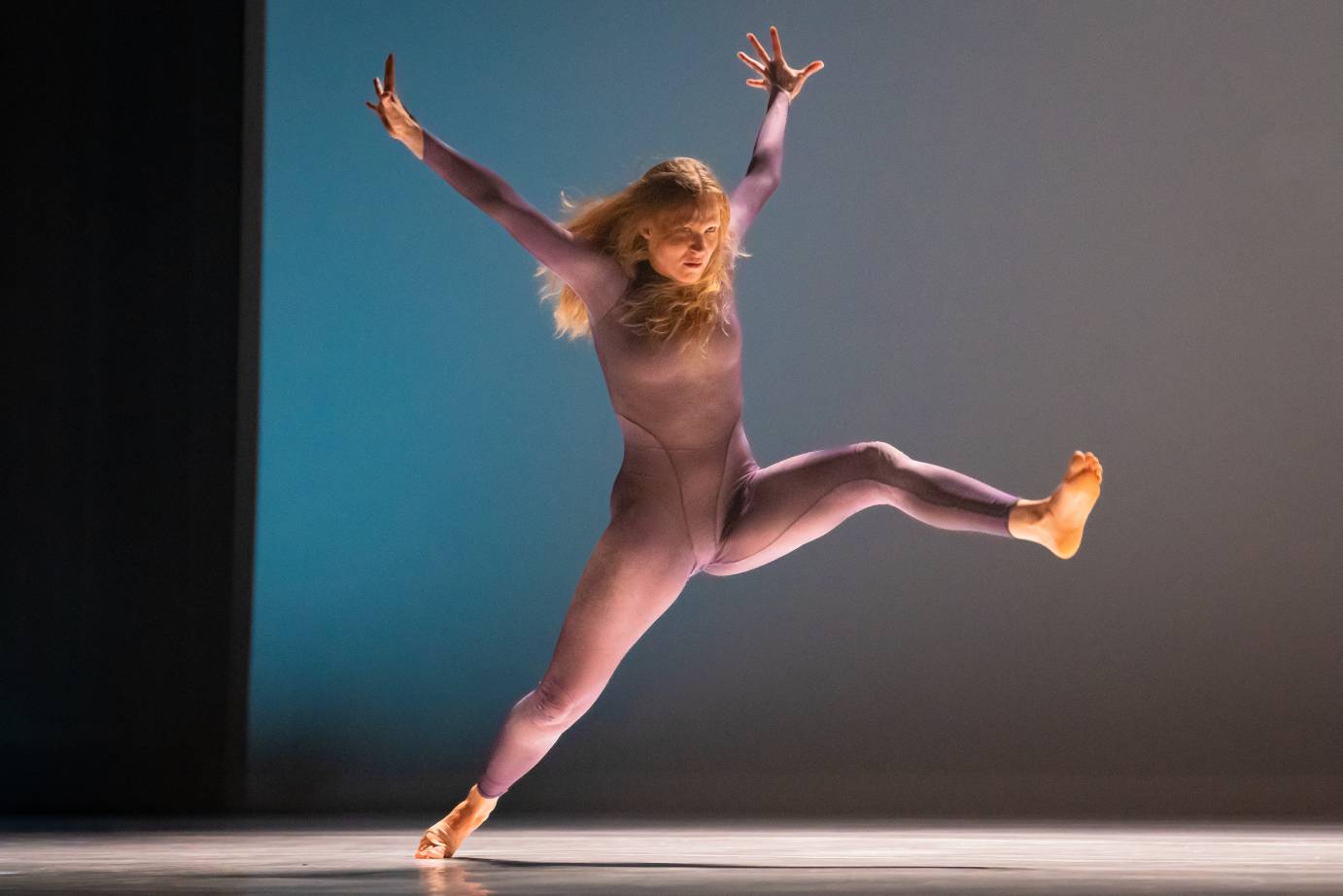 dancers in light purple unitard against a blue background dynamically kicking her leg forward as her body thrusts in the same direction arms up in a v shape to her sides and her hair flows wildly