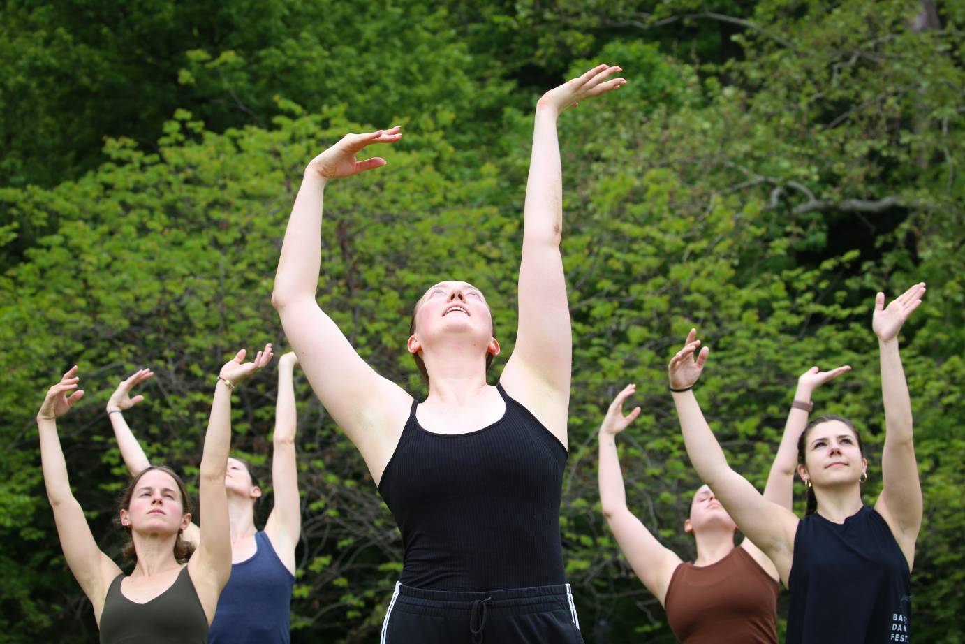 a torso view of a  company of women led by Garnet Henderson dancing in the park with their arms waving above them 