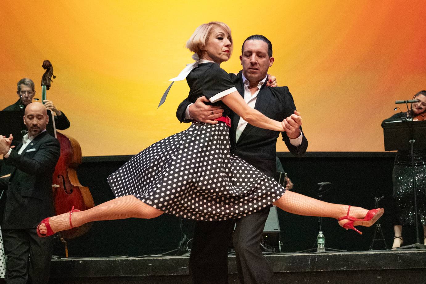 A woman in a black and white polka dotted dress and red high heels, sporting a blond bob is held horizontal around her waist by her partner.