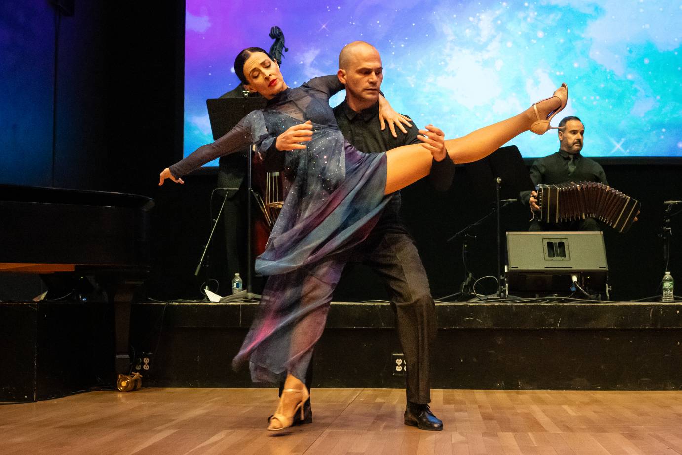 A woman in a blue gray, long-sleeved dress extends her left bare leg that is held high by her partner.
