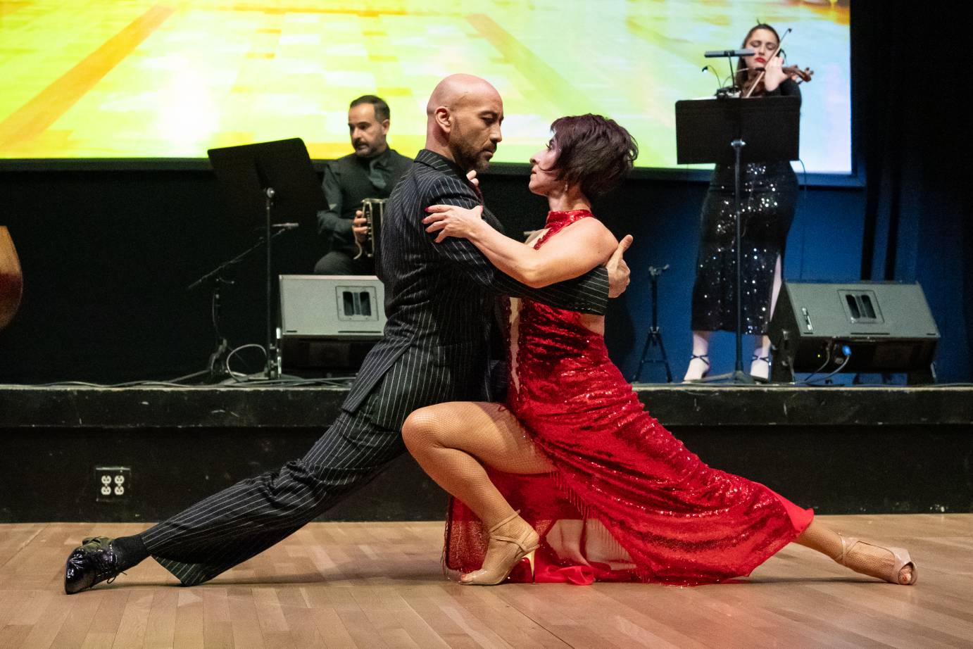 A woman in a long red dress and high heels, is clasped close to a man dressed in a black suit with vertical white stripes. The two look into each other's eyes.