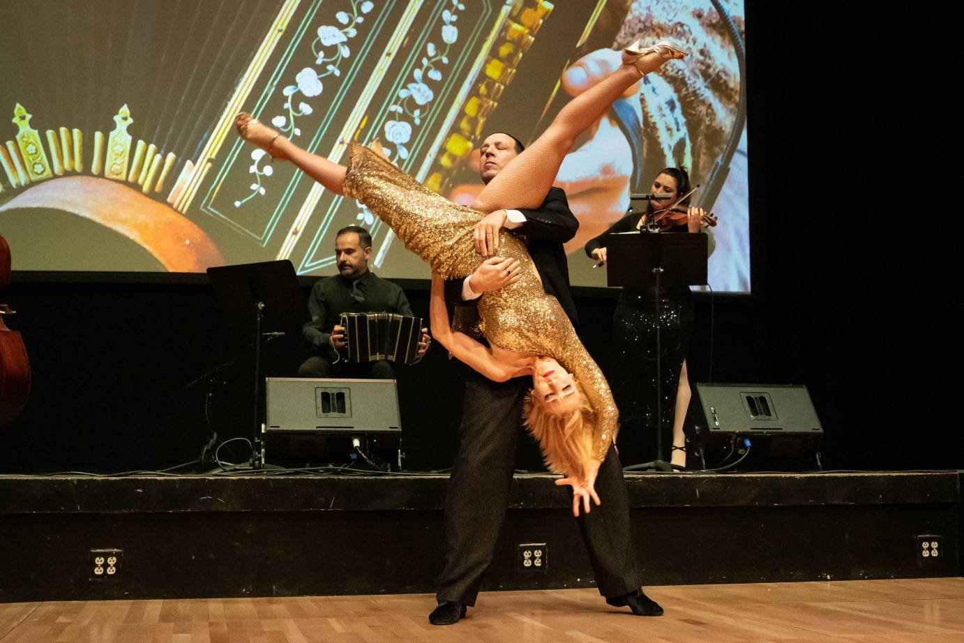 A blond haired woman in a sparkling gold dress is held upside doen with legs open by her partner who holds her dress to her body.