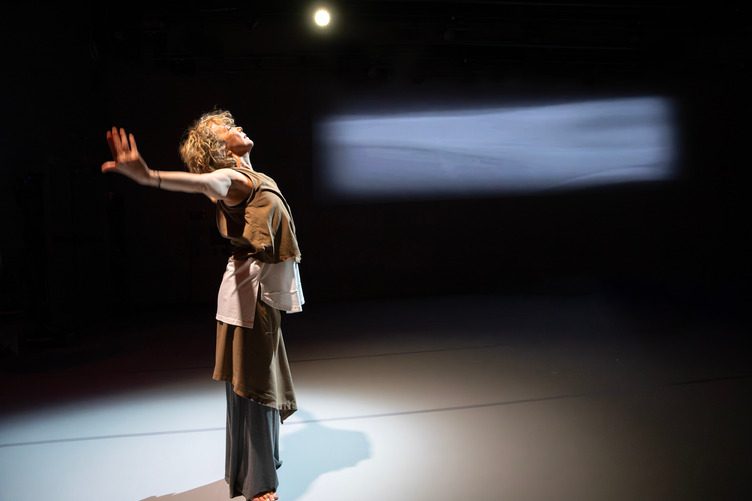 A solo woman stands with head uplifted and arms horizontal. A blued rectangular screen is behind.