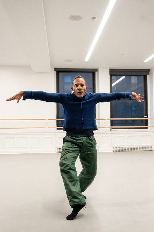 A solo man faces the camera with arms extended on the horizontal. He wears pedestrian clothes - a long sleeved blue shirt and green pants. The background are two darkened windows and a ballet barre.