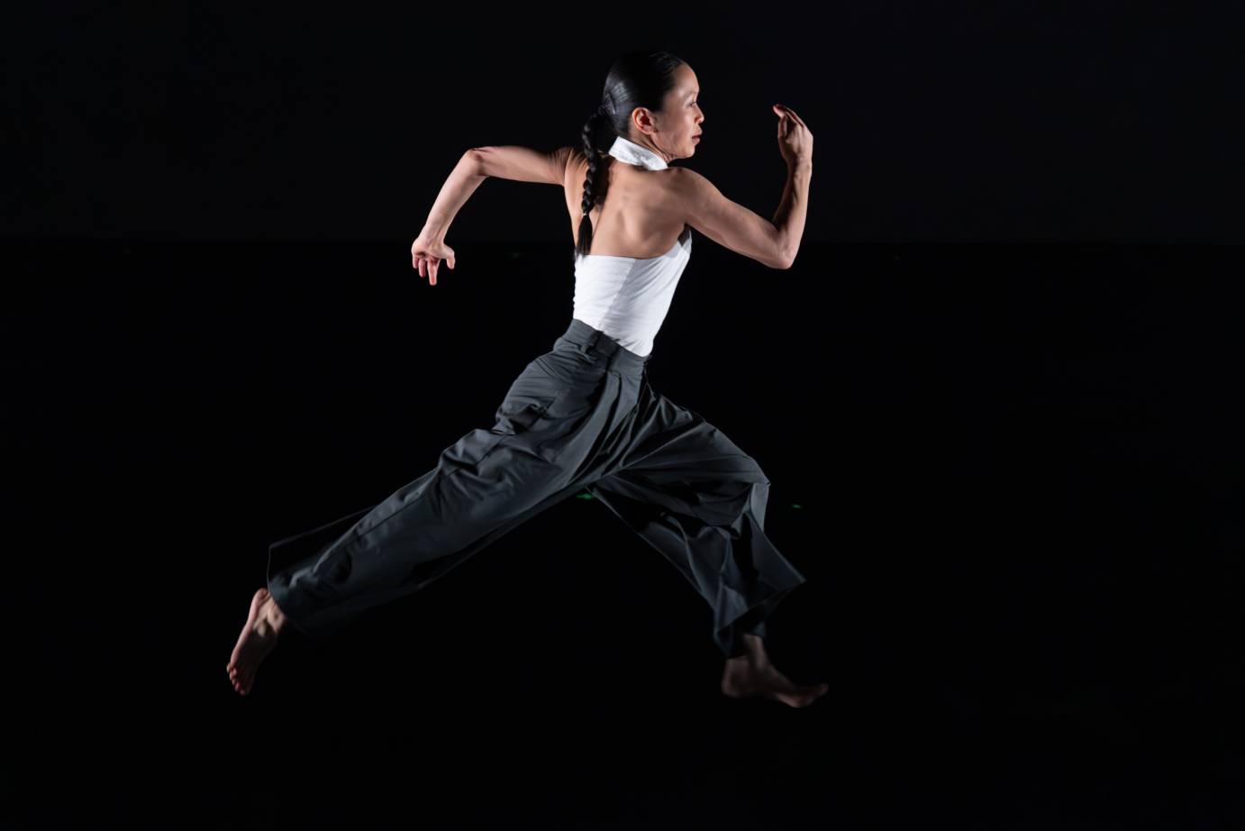 A woman in profile and dressed in a white halter top and dark pants, pumps her bare arms as she runs.