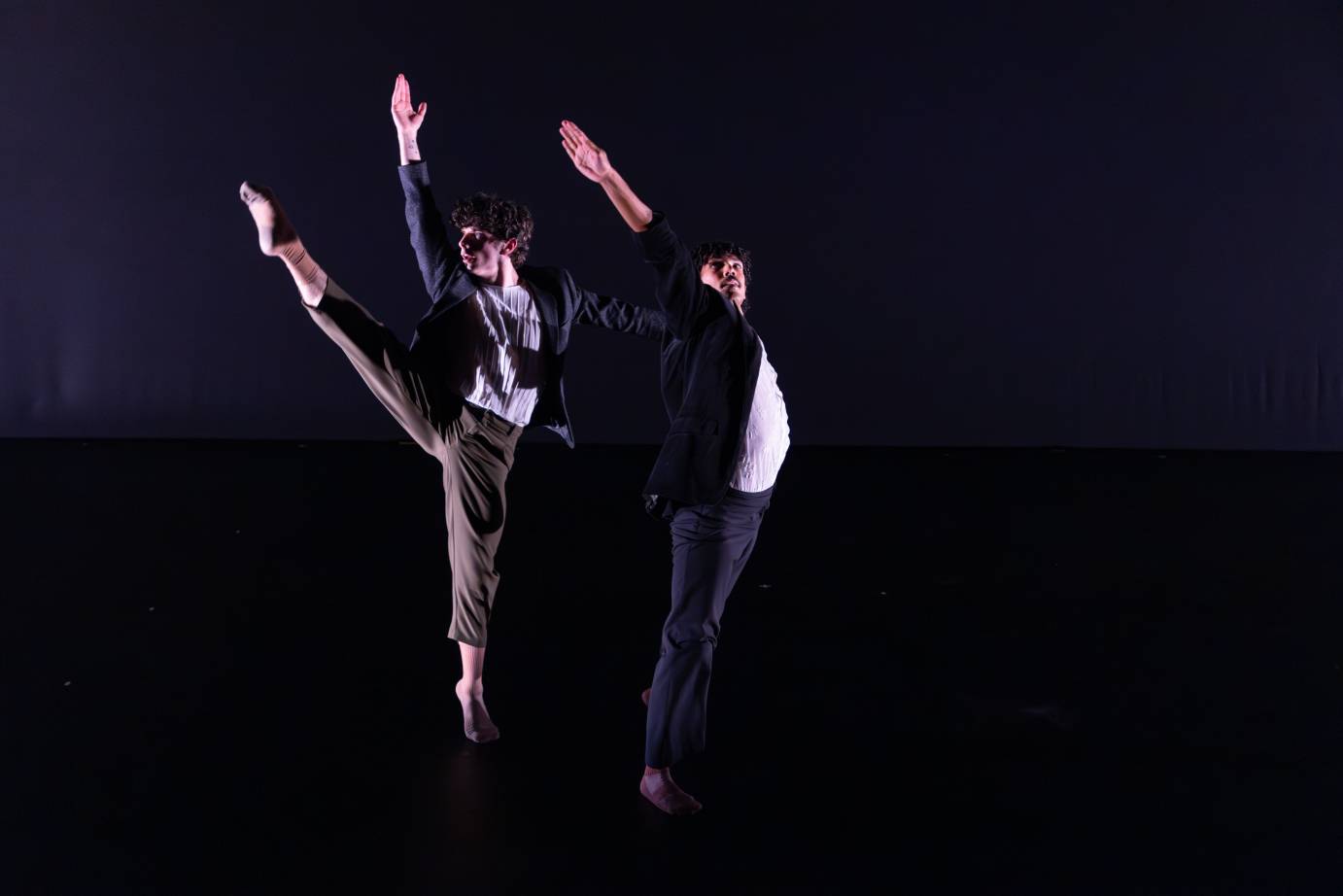 Two men dressed in everyday clothing, side by side as they jump fully extended upward. One's leg is extended far beyond his head.