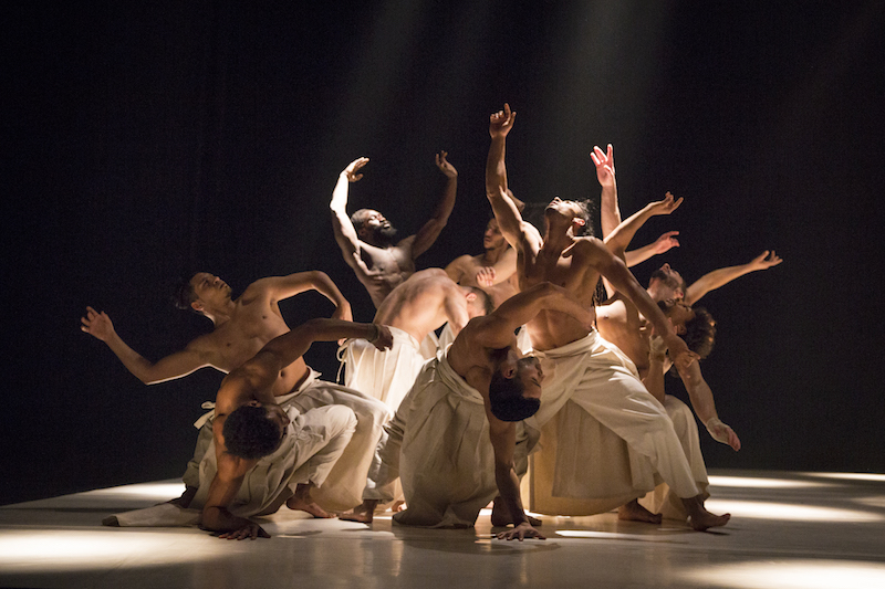 Men are in close to one another posing in a tableau. Their arms look like tentacles. 