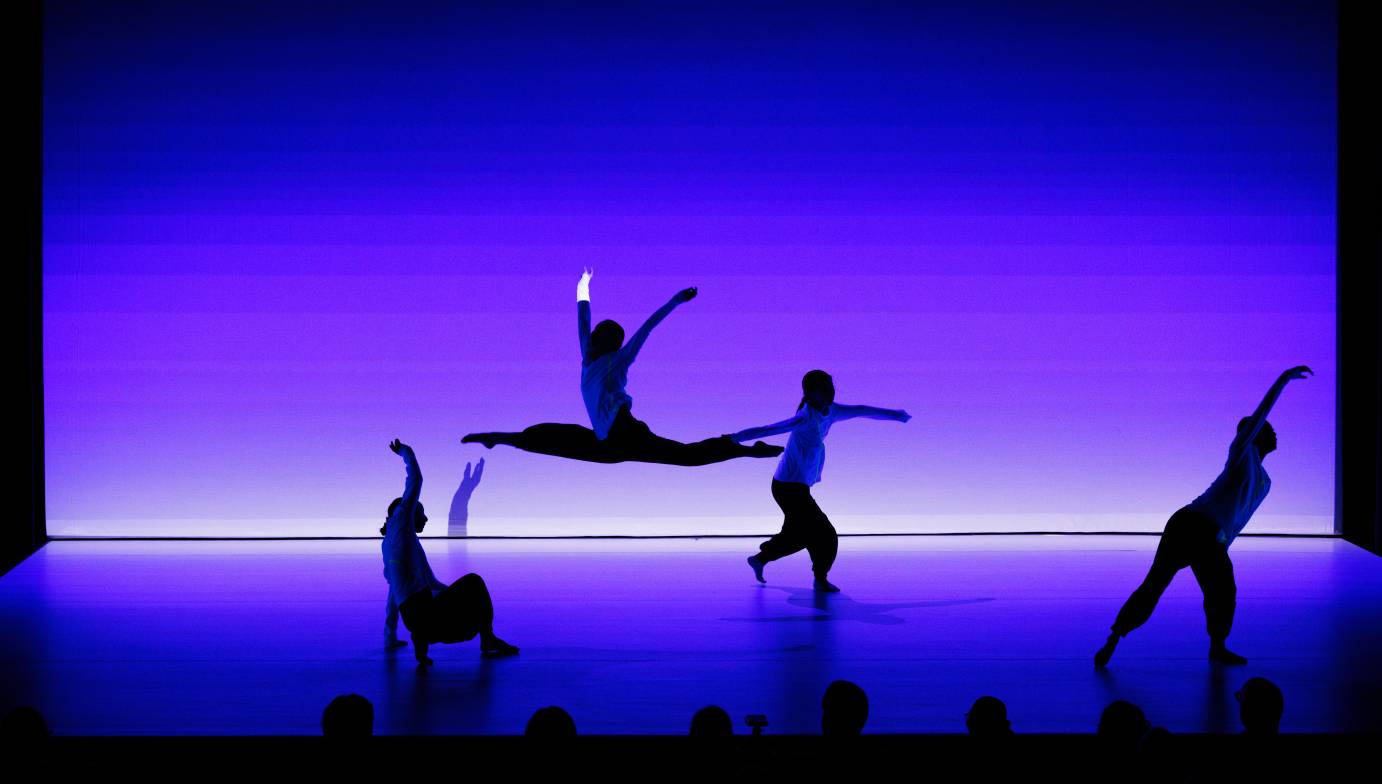 Four women dancers kneeling, reaching and jeteing against a purple background.