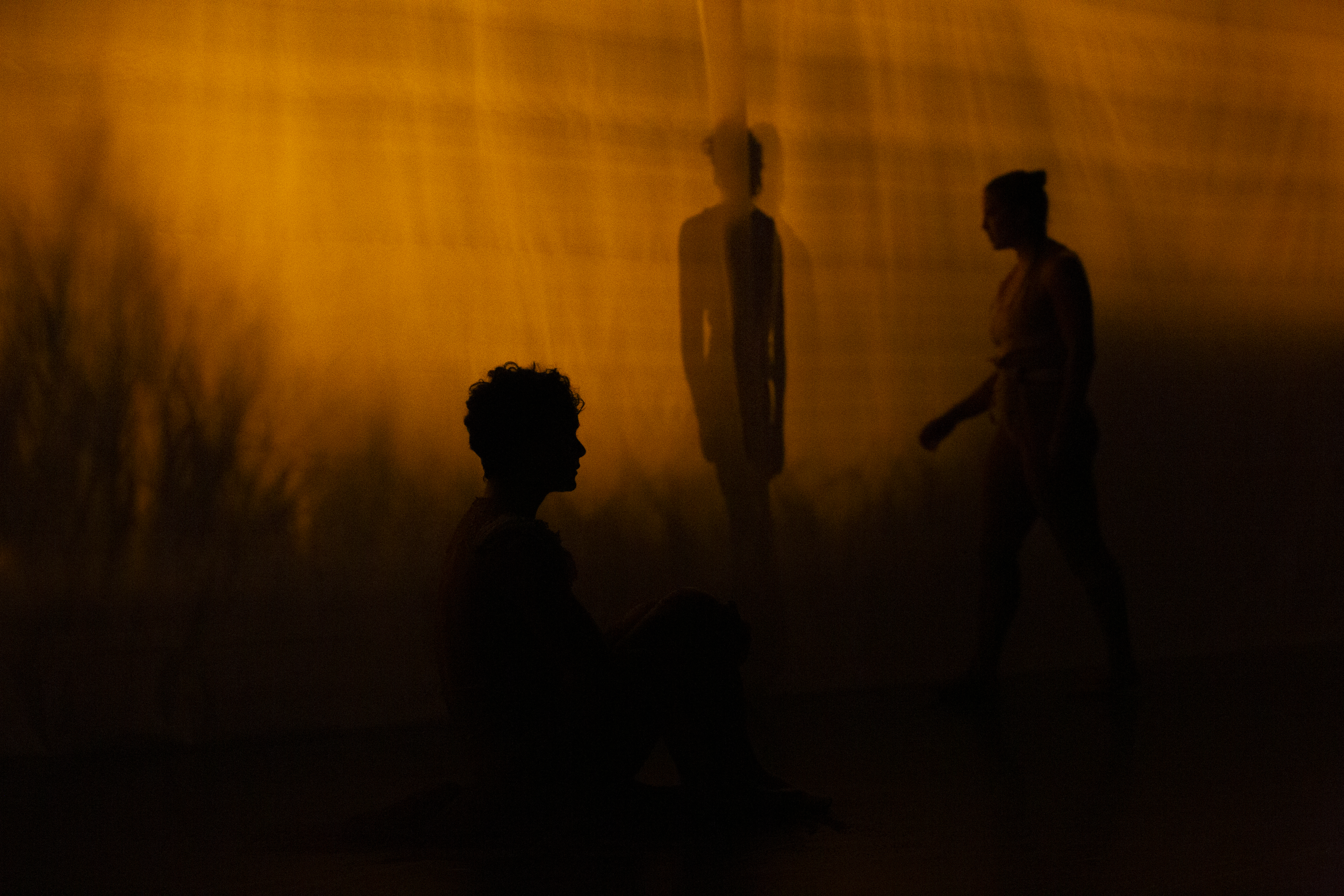Three figures standing and sitting on different levels are in silhouette against a rich amber background.