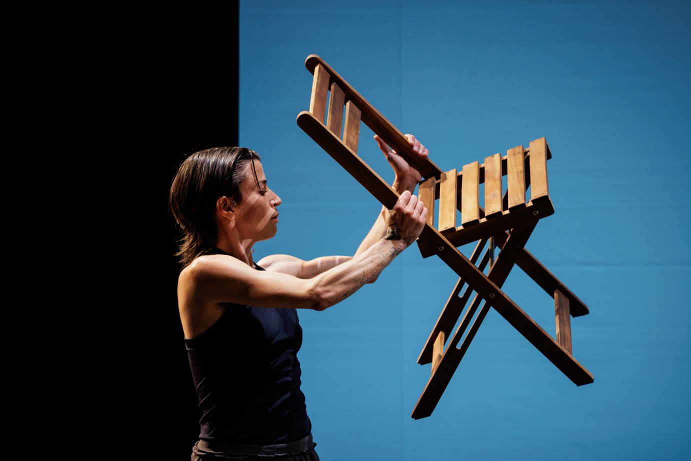 A dancer with muscular arms faces a folding chair that they lifted.