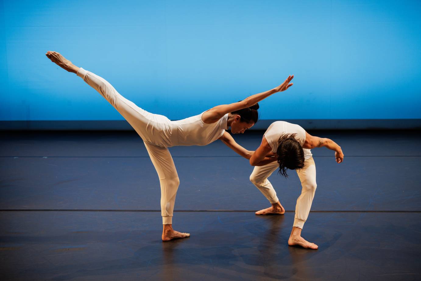 Two dancers, both dressed in white shirts and leggings, one with a bun at the top of her head, standing in an extremely arched arabesque contrasts with the other who is folded forward the top of their head to the audience.