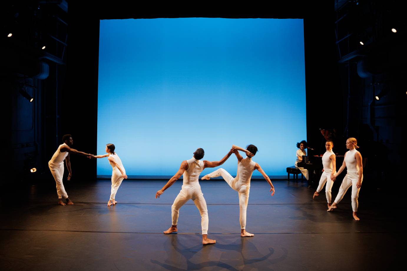 Three pairs of dancers spaced evenly between the dancers and the pairs situated in front of a baby grand piano and pianist. All the pairs are dressed in white and all on their feet.