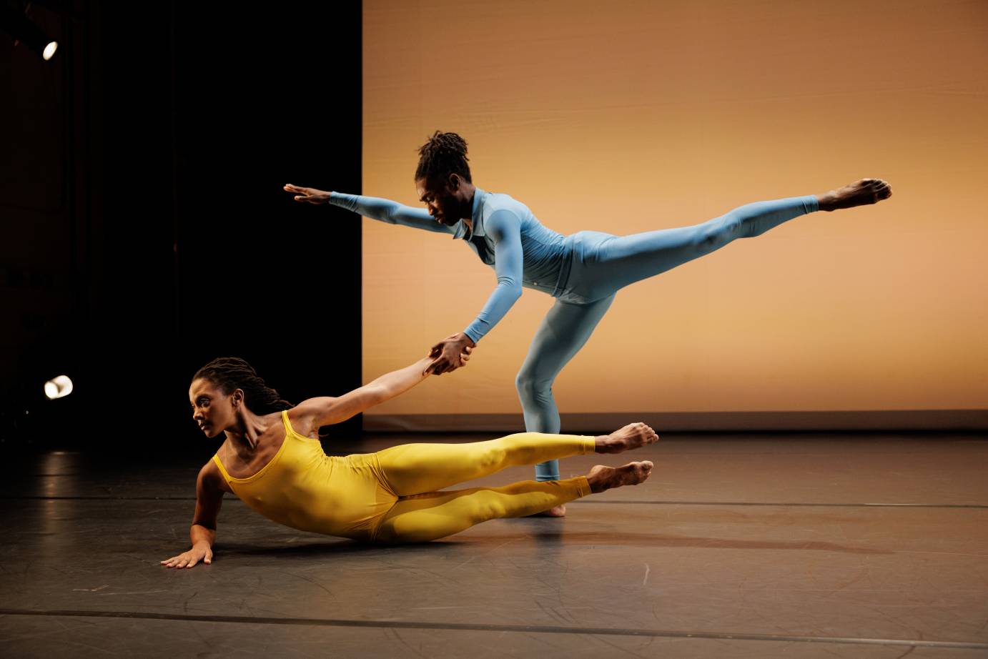 Two performers - one in a blue unitard in arabesque holds the hand of a dancer in a yellow unitard stretched long on her side.