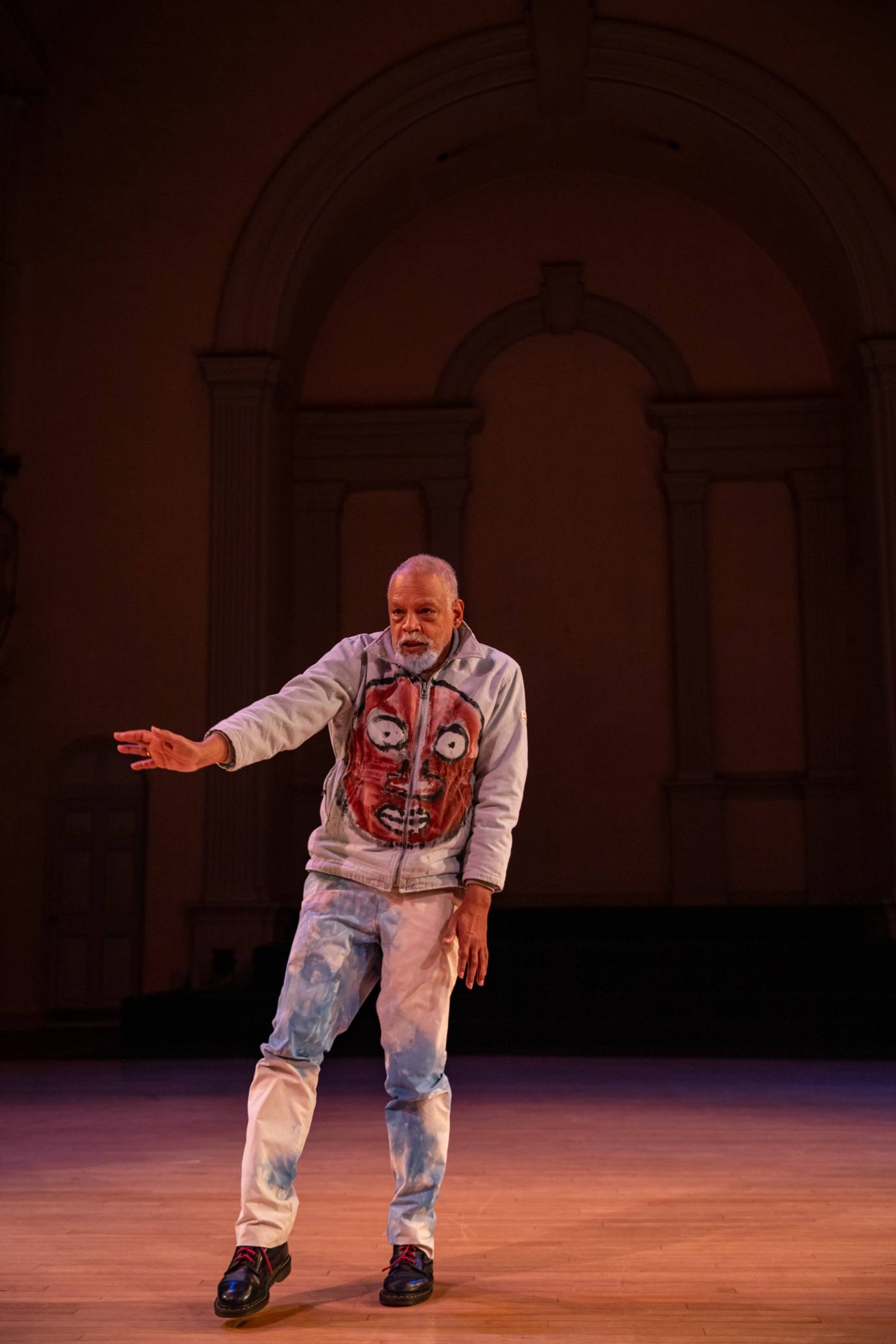 A solo man faces forward gesturing with his right hand. His hoodie portrays a round, dark face with wide eyes.