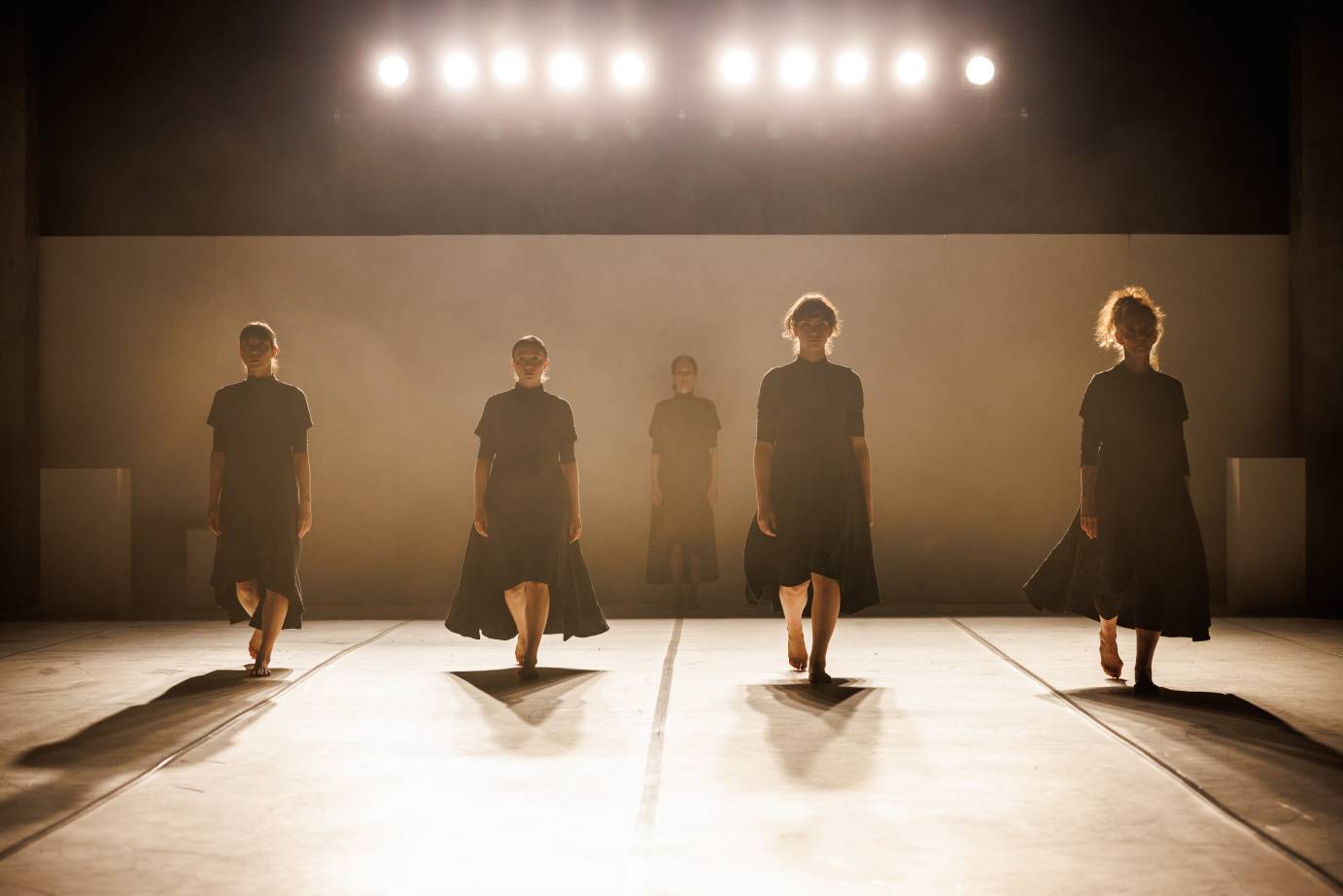 Five women in black mid calf dresses face downstage backlit by ten strong lights. Their hair aglow.
