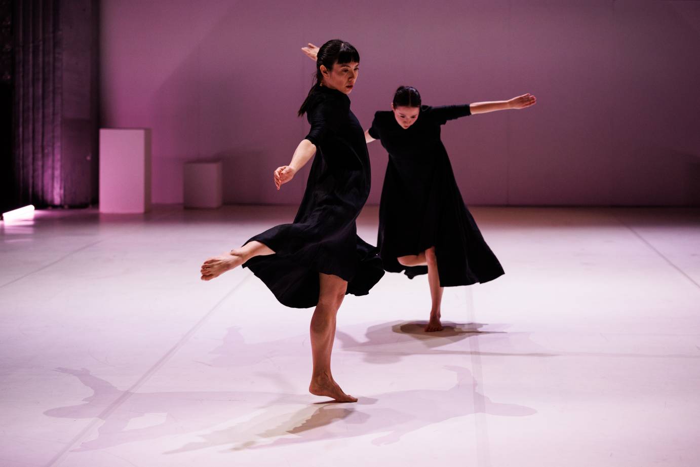 Two women dressed in black flowing dress with sleeves to the elbows toss arms and rise slightly off the floor, one leg behind.