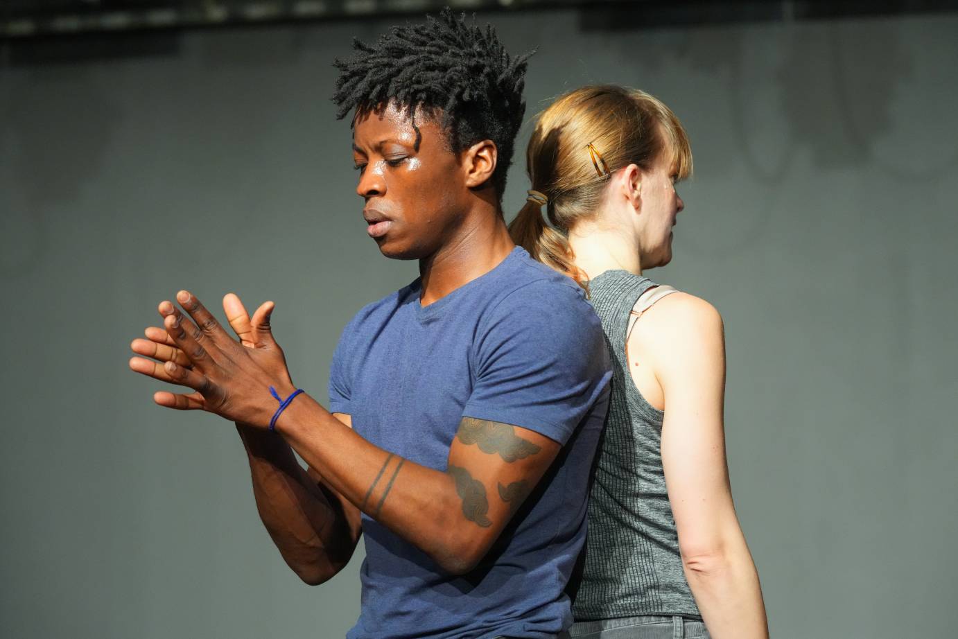 Mufutau Yusuf and  Jamie Scott  stand back to back. Jamie's face is hidden but she appears to be speaking.Mufutau glances at his hands while rubbing them together. We notice dark tattoos on his arms that look like birds. 