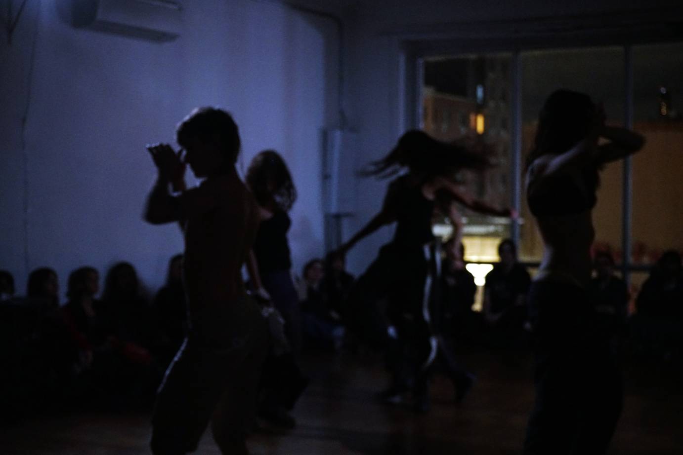 The silhouettes of performers dancing. The background is bluish and the audience is seated watching.