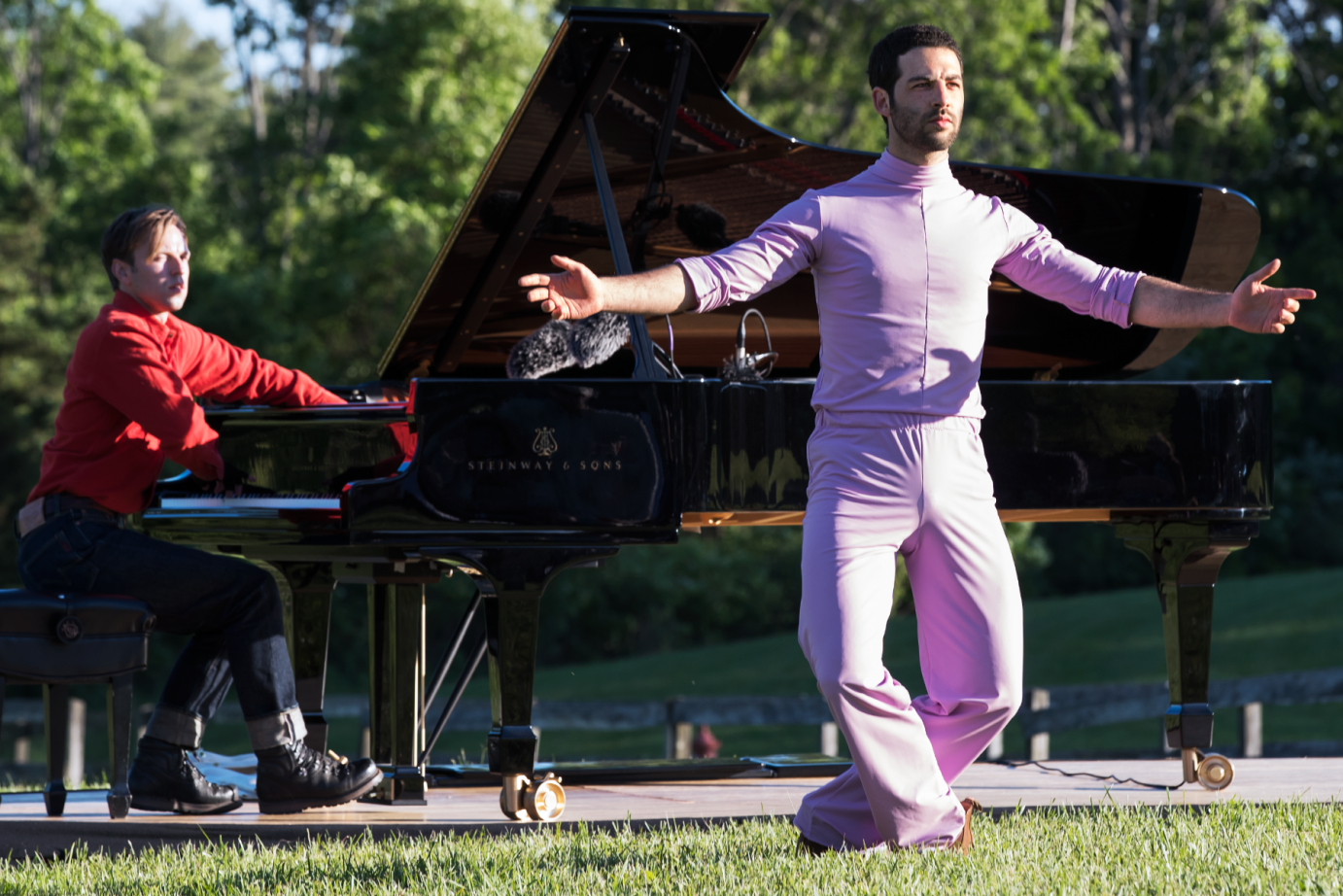 Or Schraiber dressed in a silky pink turtleneck neck with matching pants faces the audience in a balletic knee band in the  foreground, while behind him Hunter Noak manipulates his piano’s internal hammers/