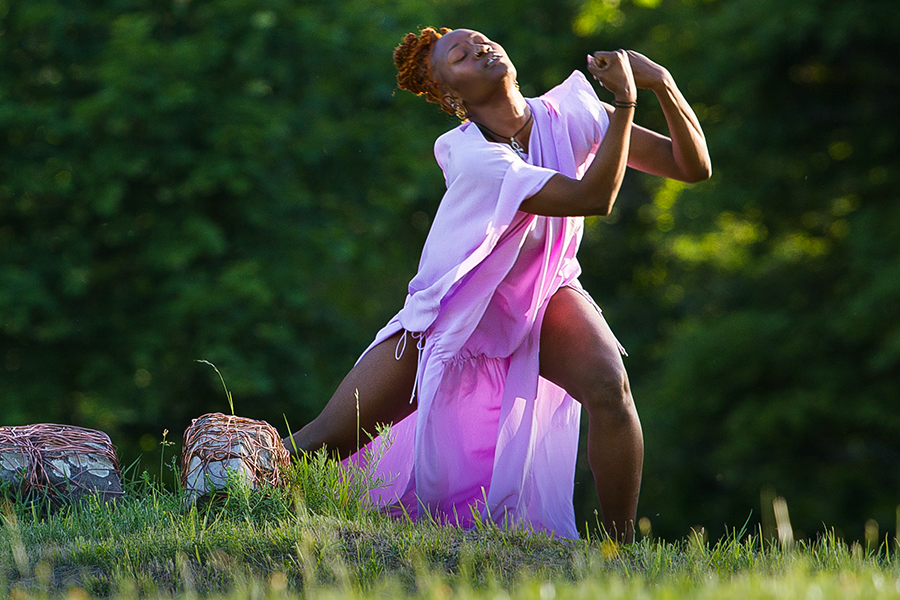 Coral Dolphin, the Black woman in the flowing pink tunic, stands in the grass, in front of largish rocks, holding her hands in front of her leaning towards one bent leg while the other is planted straight behind her. She appears to be pleading. 
