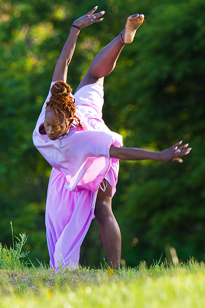 Coral Dolphin, the black woman in silk pink tunic, appears to be diving towards the ground. One leg is extended behind her as her body leans forward and her eyes look towards the ground. 