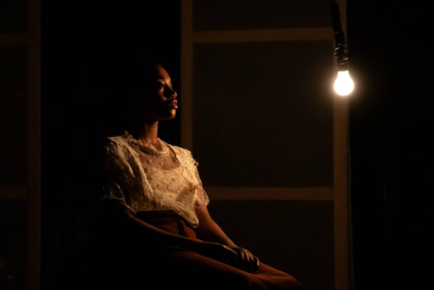 A woman in a polka-dotted blouse and rust-colored skirt sits in a chair looking at a bare bulb hanging in front of her face.