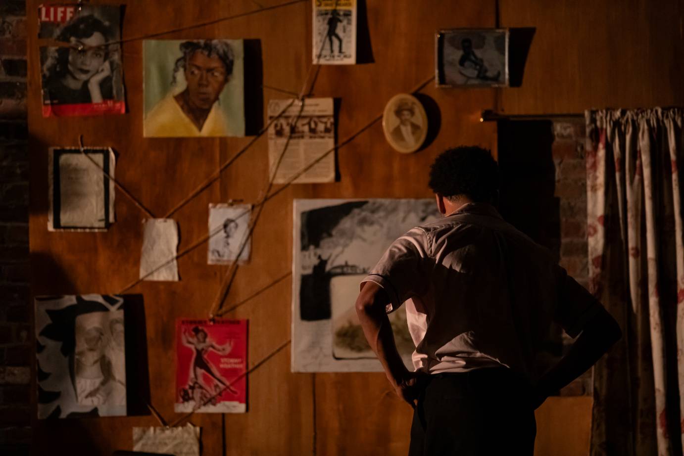 A wall of portraits hang on a reddish wall. A man with his back to the audience stands at the edge of the space.