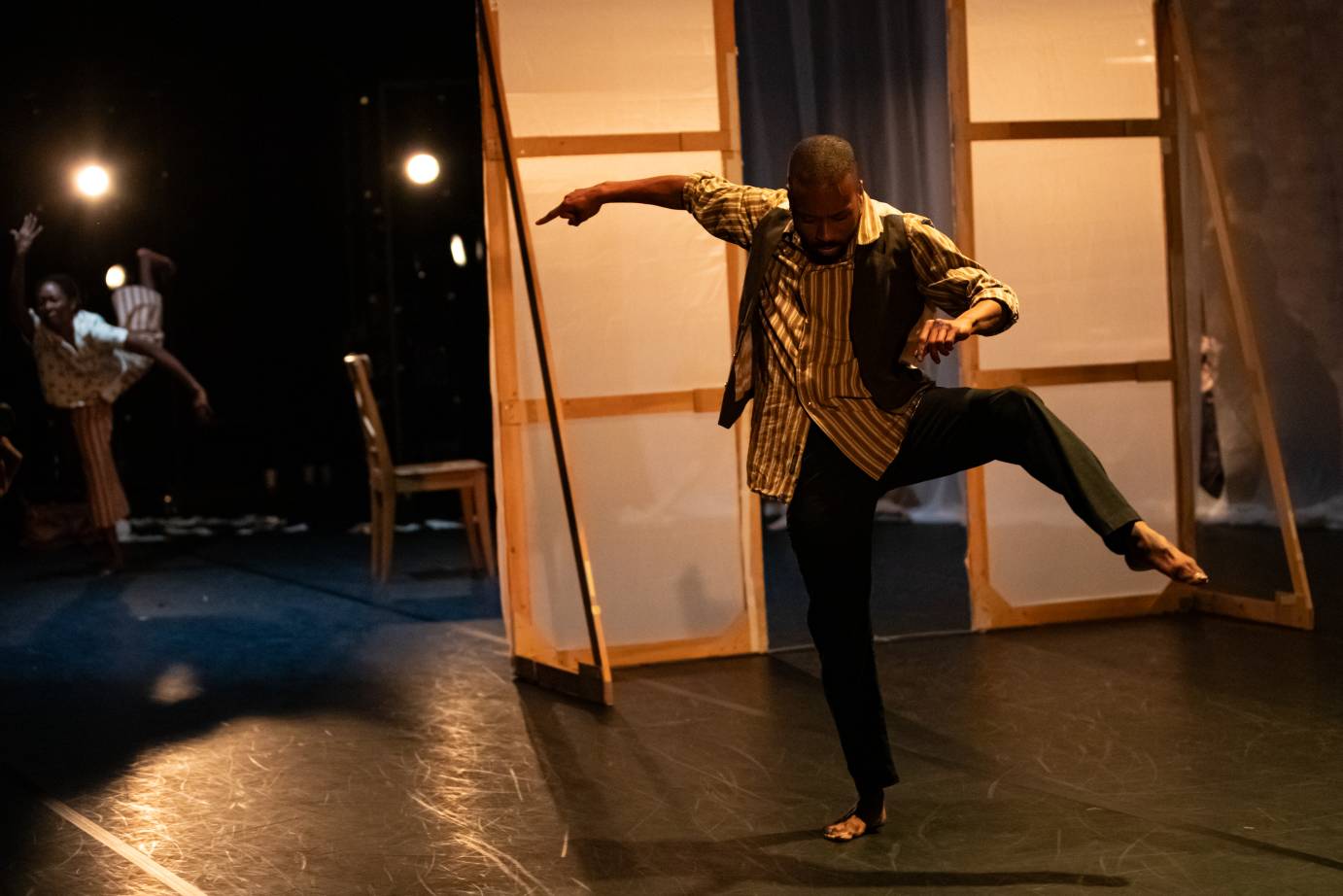 A man standing on his right leg with the left leg bent and lifted, arms on the horizontal, looks down. He is dressed in a striped shirt, vest, and everyday pants.He dances in front of an opaque screen.