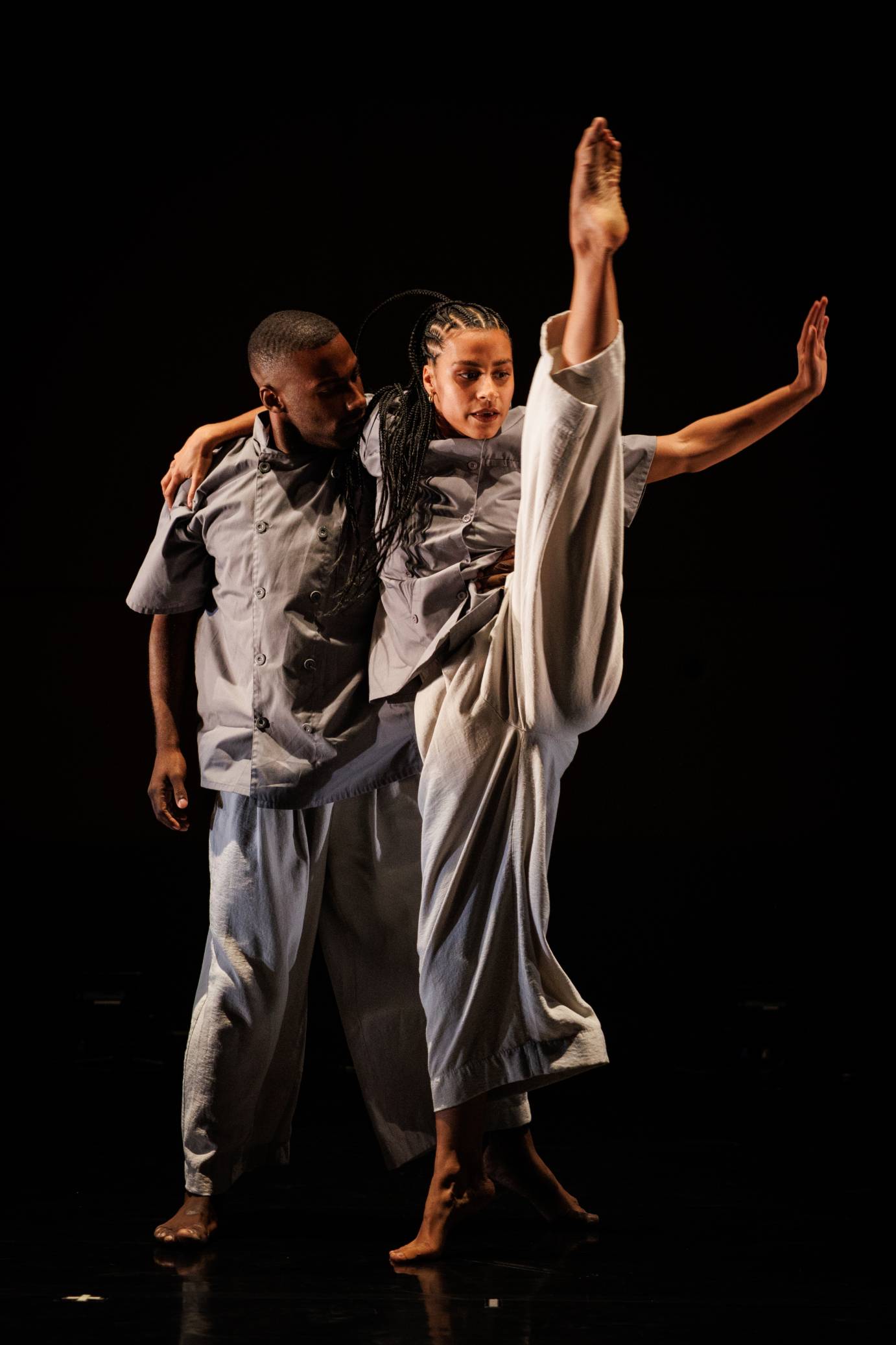 A woman slings her arm over a man's shoulder as he looks at her face while she extends her leg far above her head, her left arm extended with a bent elbow.