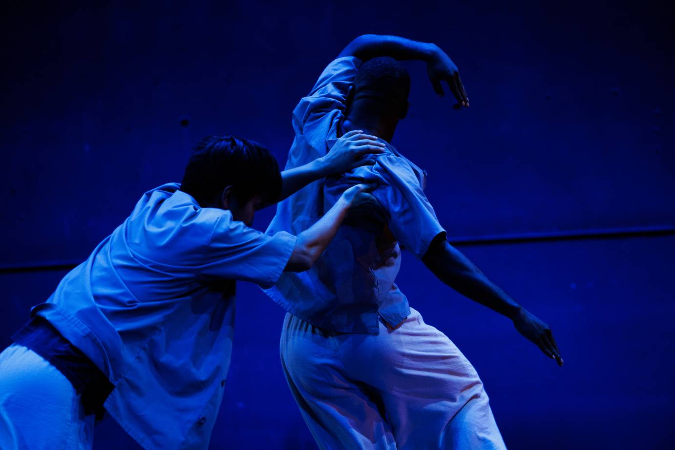 Two men are bathed in blue light, backs to the audience, with the man behind holds onto to the man in front.