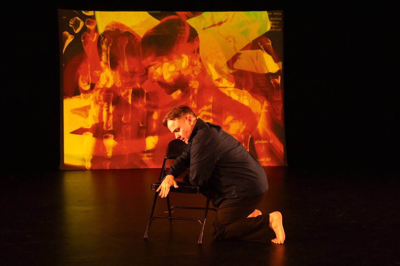 A man dressed in black in front of a bright red and yellow painting projection, half kneels while holding onto a black folding chair.