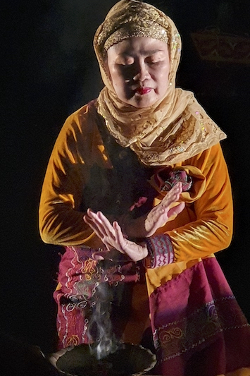 Portri Ranka Manis a Filipino woman,head wrapped in yellow bejeweled scarf, wearing a mustard colored dress, and a mostly red wrap around her waist, sits before a bowl of incense, arms crossed and palms open in front of her body. her eyes are closed in meditation or prayer