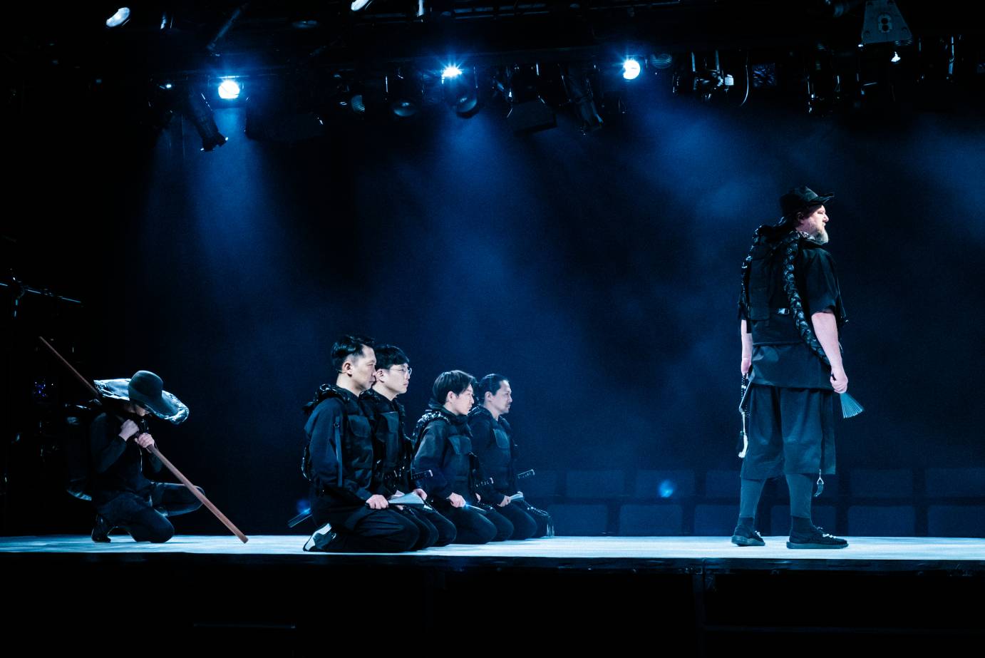 Six male performers are arranged with a soloist in front of four kneeling performers dressed in black followed by a performer bending forward with a long stick.