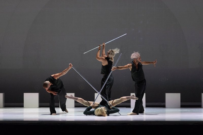 Dancers in vests and slacks hold metal poles. One dancer lays on their back with their legs splayed. The others stand in mid-movement.