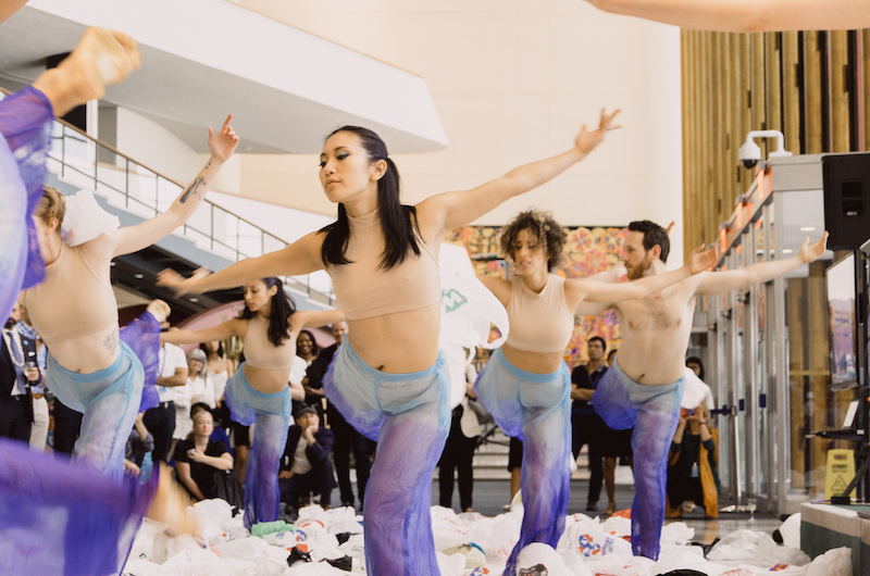 Dancers in purple tie dye pants and nude crop tops strike a pose with one leg bent behind them in a white lobby. Plastic bags are strewn about their feet. A crowd watches in the background.