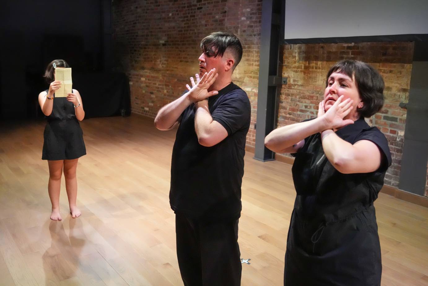 A girl in black dress holds a paper in front of her face. A man and woman (her parents) cross their hands and press them into their cheeks
