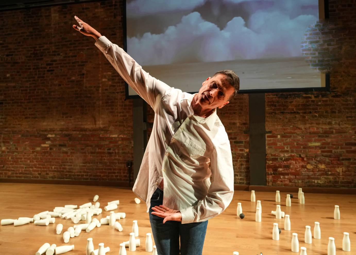 Man in white dress shirt lifts his right arm on the diagonal, his left arm is curled close to his pelvis. The background includes white milk bottles strewn on the floor and an image of an exploding bomb on the screen behind.