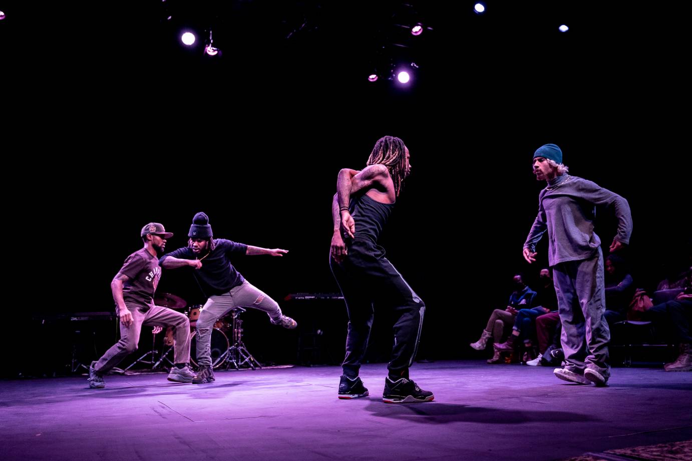 2 Pairs of male Black dancers seem to speak to one another using poses. The man in the front bends his arm backwards in an almost contorted position as his partner stares at him almost defiantly, like a stiff robot. The other couple int the back contact one another shoulder to elbow.