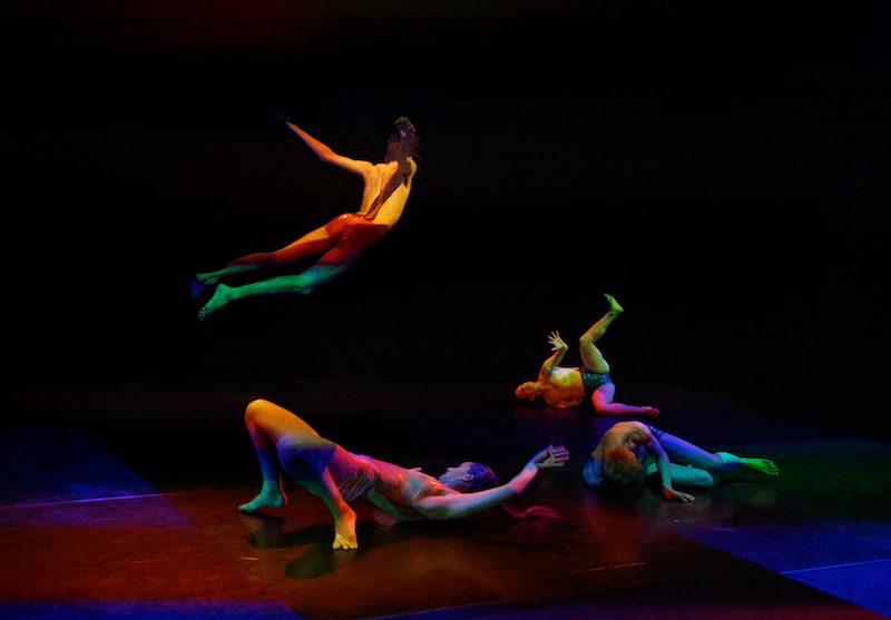 Five dancers in PVC shorts writhe on the floor. One dancer is airborne like Superman/Superwoman.