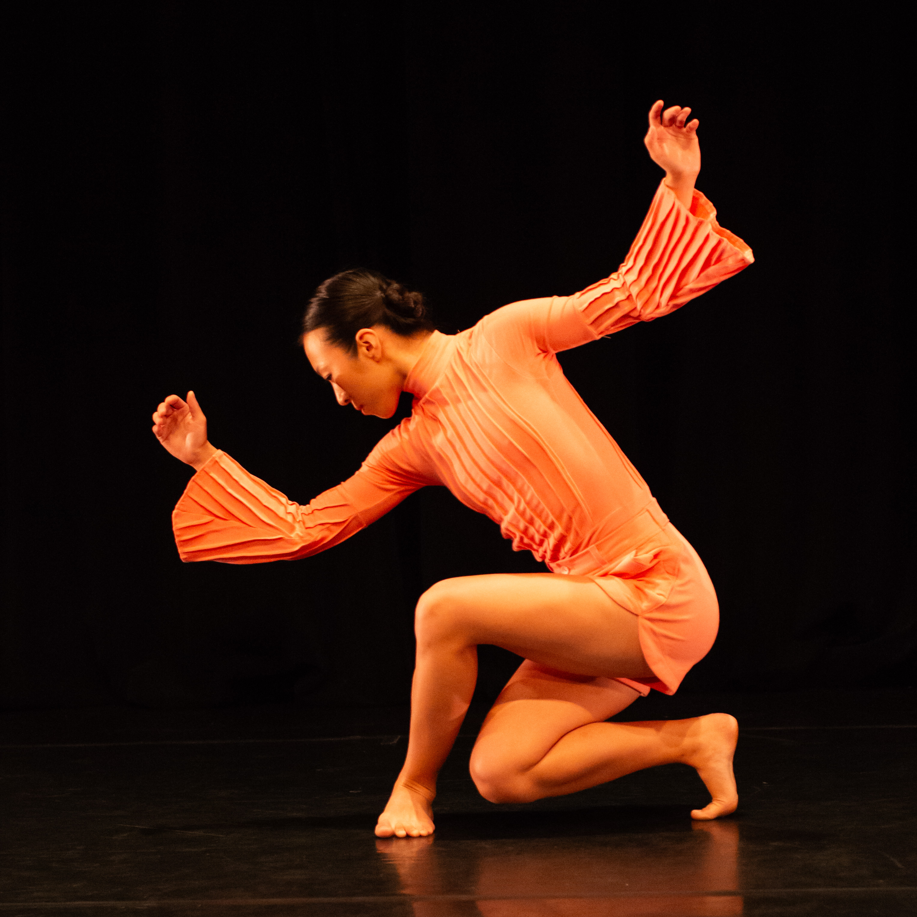 a woman in profile bows regally she wears a beautiful orange costume with a textured bodice connected to short pants 