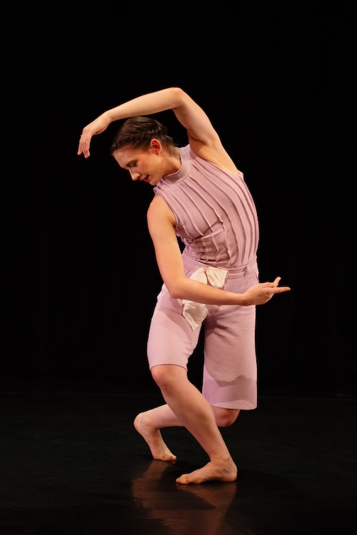 a studio shot of a woman in profile wearing a light lilac costume ... one arm creates a semi-cirecle above her , the other a semi circle below her torso. Her top has no sleeves and has textured stripes vertical placed on it, she wears shorts that extend toward her knees. a white hankerchief  is tucked into the waist of her pants.