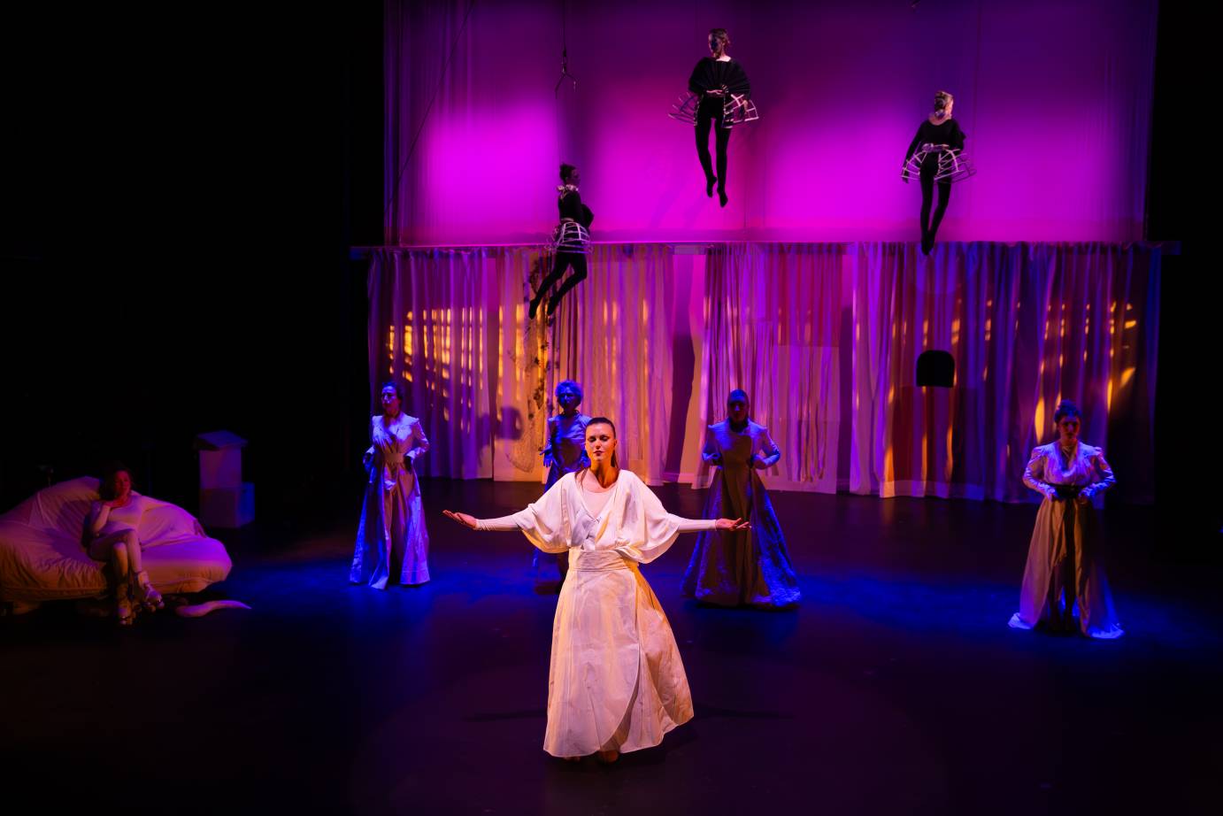 A woman in a long white dress stands forward of four women upstage. Her arms are gesturing to the side of her body and her face is placid.