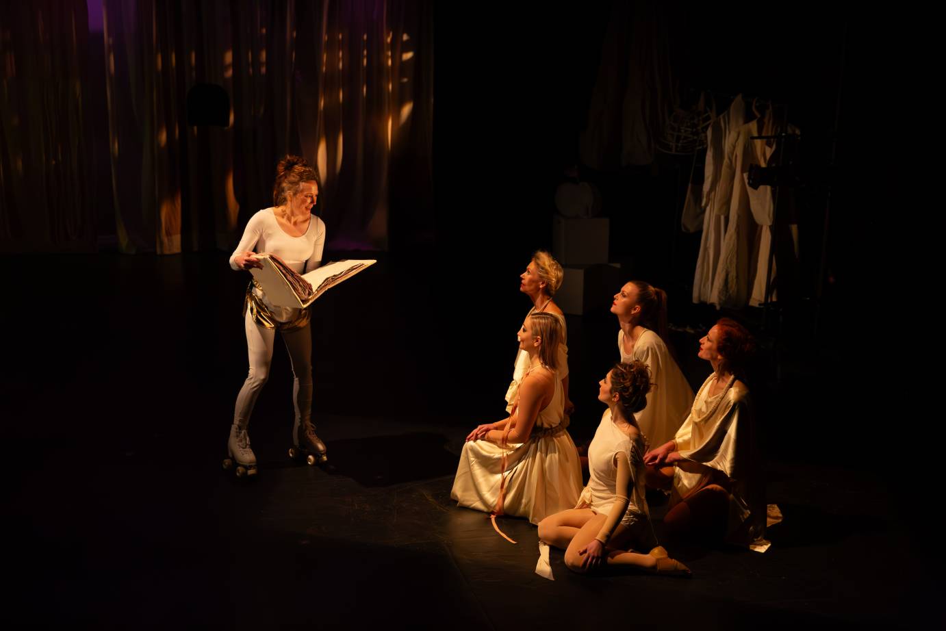 A woman dressed in a white shirt, gold shorts and skates, holds a large album in both hands as she reads to the five women accolytes with faces upturned to her.