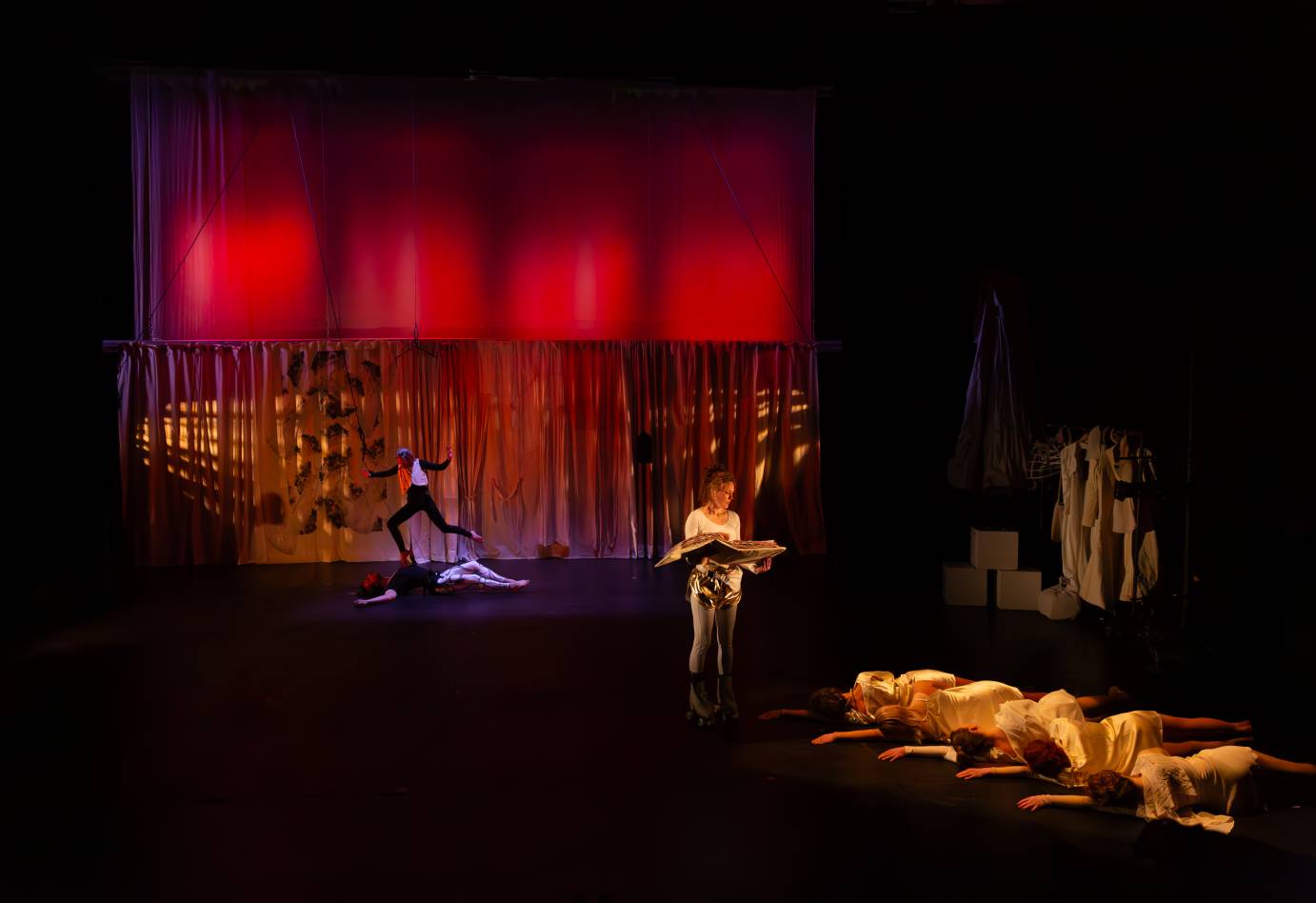 The woman dressed in skates continues to read from the album in her hands to the five women now lying on their sides. Upstage is a woman aerialist tiptoeing on the chest of a man, also an aerialist, who has levitated a few inches off the floor.  
