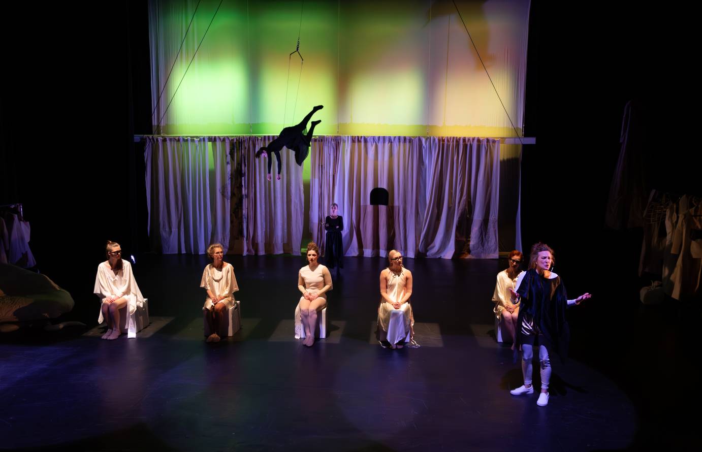 The Narrator, now in sneakers and a black tunic stands gesturing, her face impassioned, in front of five seated women wearing glasses and white garments. A woman aerialist in a long black dress hover upstage upside down.