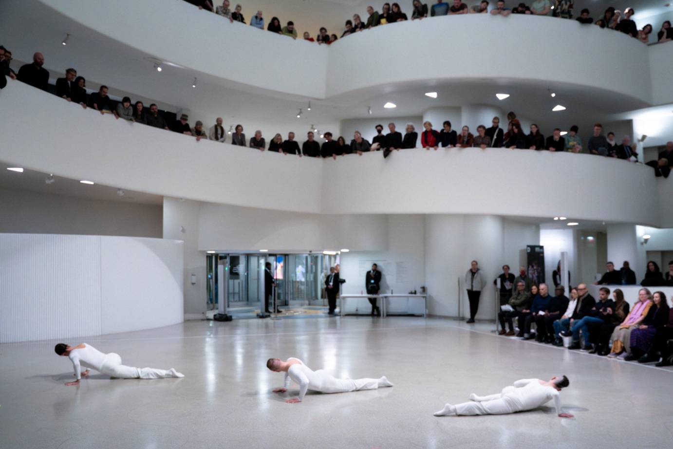 Three men, costumed in white pants and long shirts, stretch their bodies along the floor.