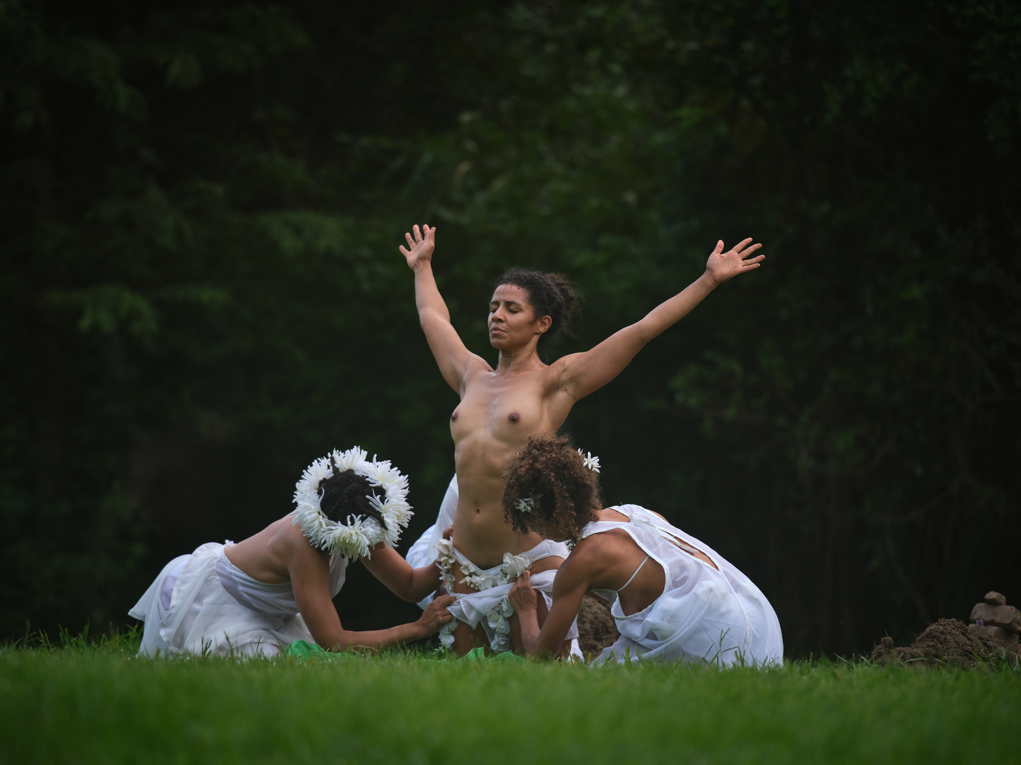 Outdoors, three dancers are positioned on a lush green lawn against a dark, forested background. One dancer stands topless at center, arms stretched upward in a powerful gesture, eyes closed. Two other dancers kneel at her sides, dressed in flowing white garments and floral headpieces, appearing to assist or adorn the central figure.