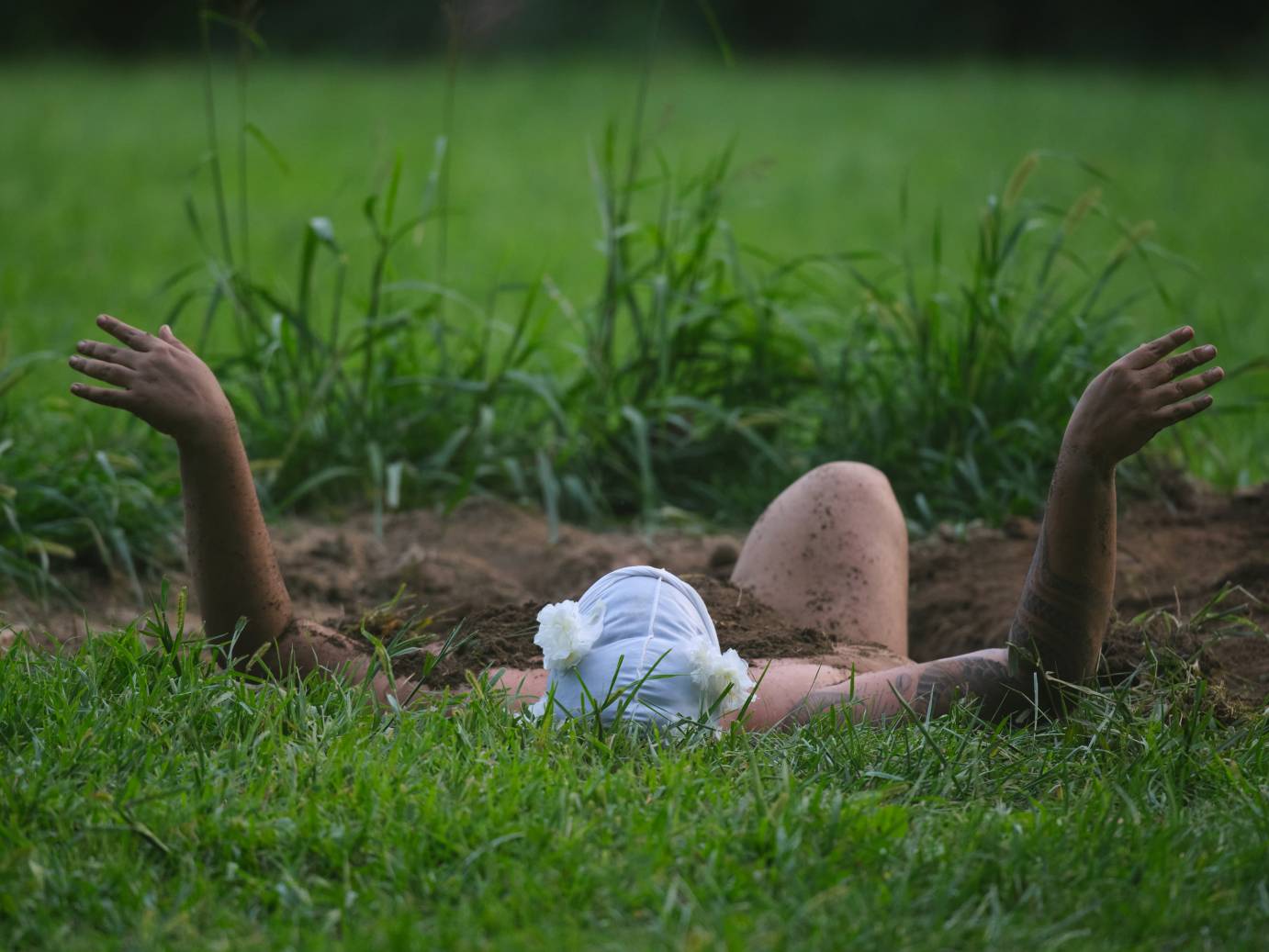 Outdoors, a dancer is pictured from behind, with a white du-rag on their head. Their arms are visible from the forearms up, with their upper arms, torso and left leg submerged in a wide hole in the ground. The dark brown of the soil where the land is open contrasts bright green lush flora.